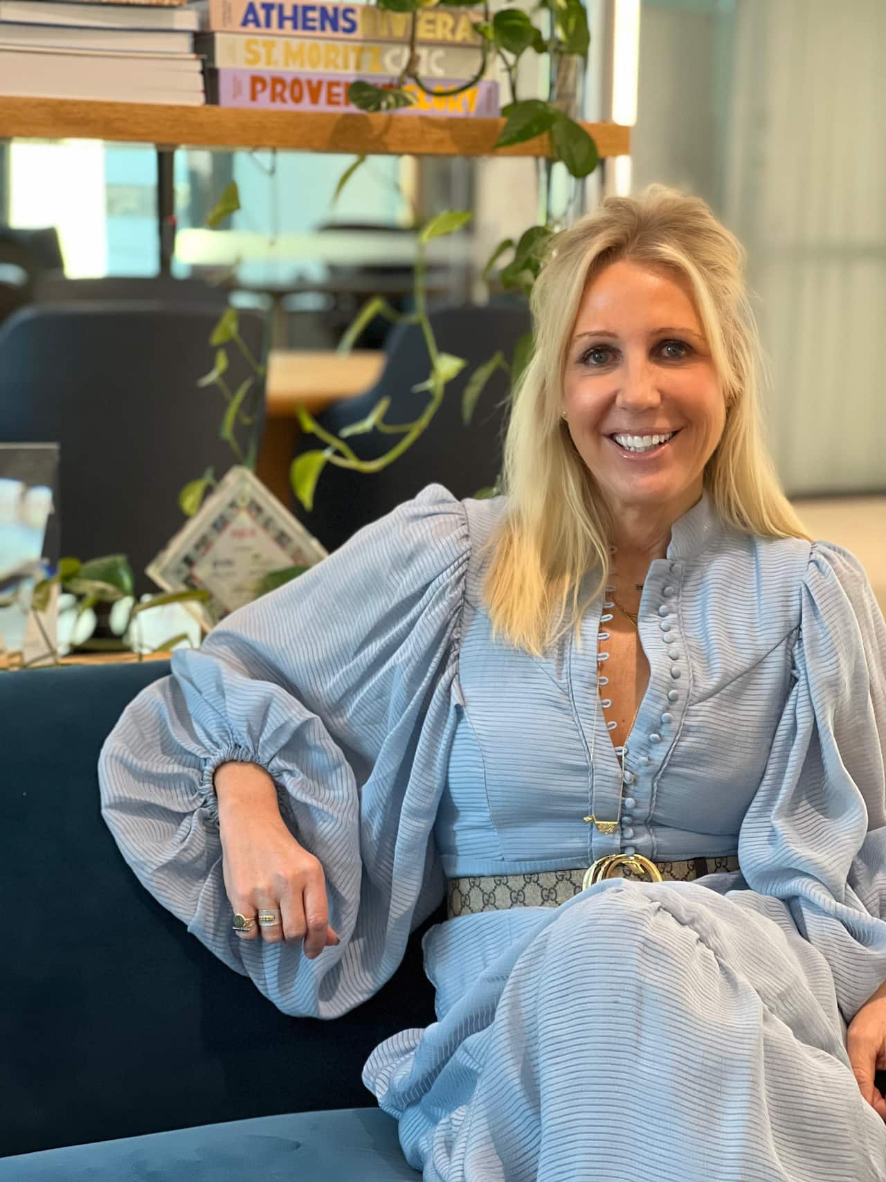 a blonde woman wearing a blue dress smiles at the camera while sitting on a couch. 