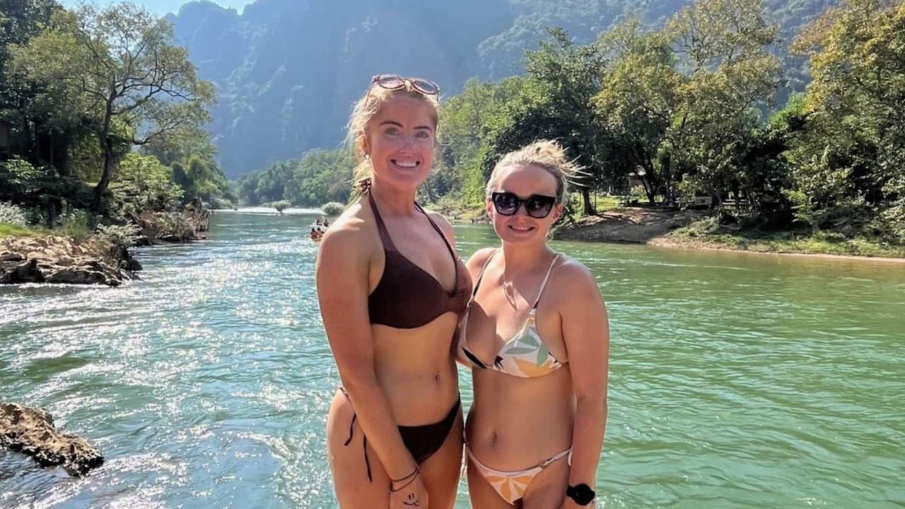 two young blonde woman smile in bikinis in front of a blue lagoon