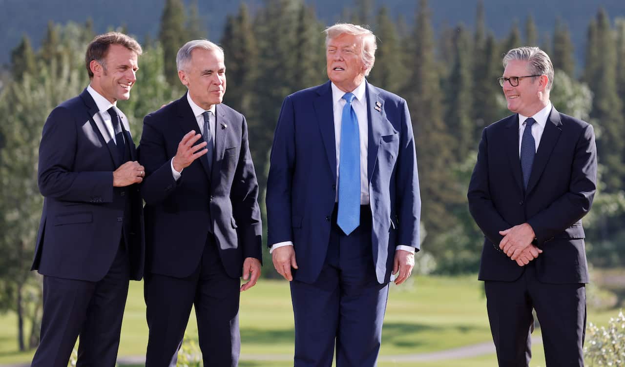 Four men in suits standing in an outside area in front of a row of trees.