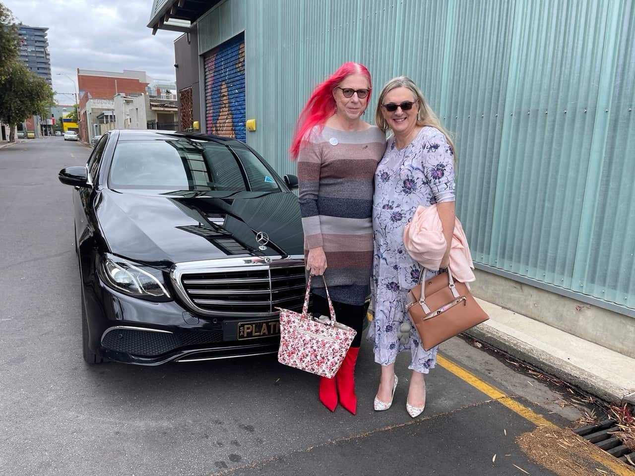 Two women carrying handbags stand in front of a blacked parked car. 