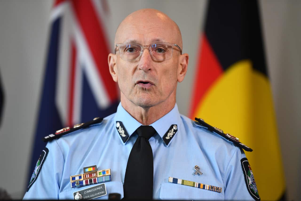 A bald man with glasses dressed in a police uniform stands in front of the Australian and Aboriginal flags.
