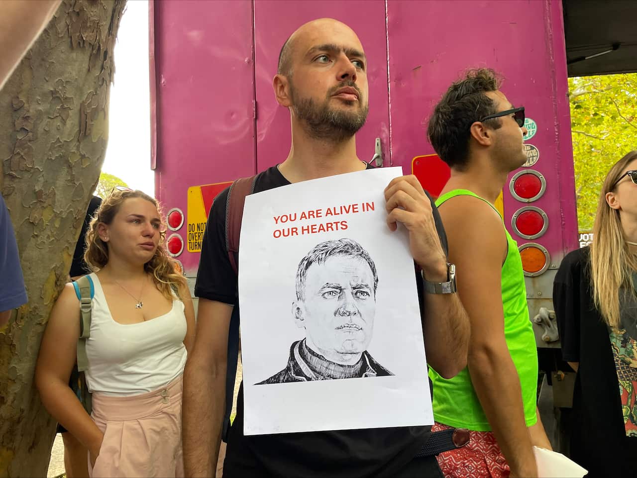 A man holds a placard with a photo of Alexei Navalny with two people in the background.