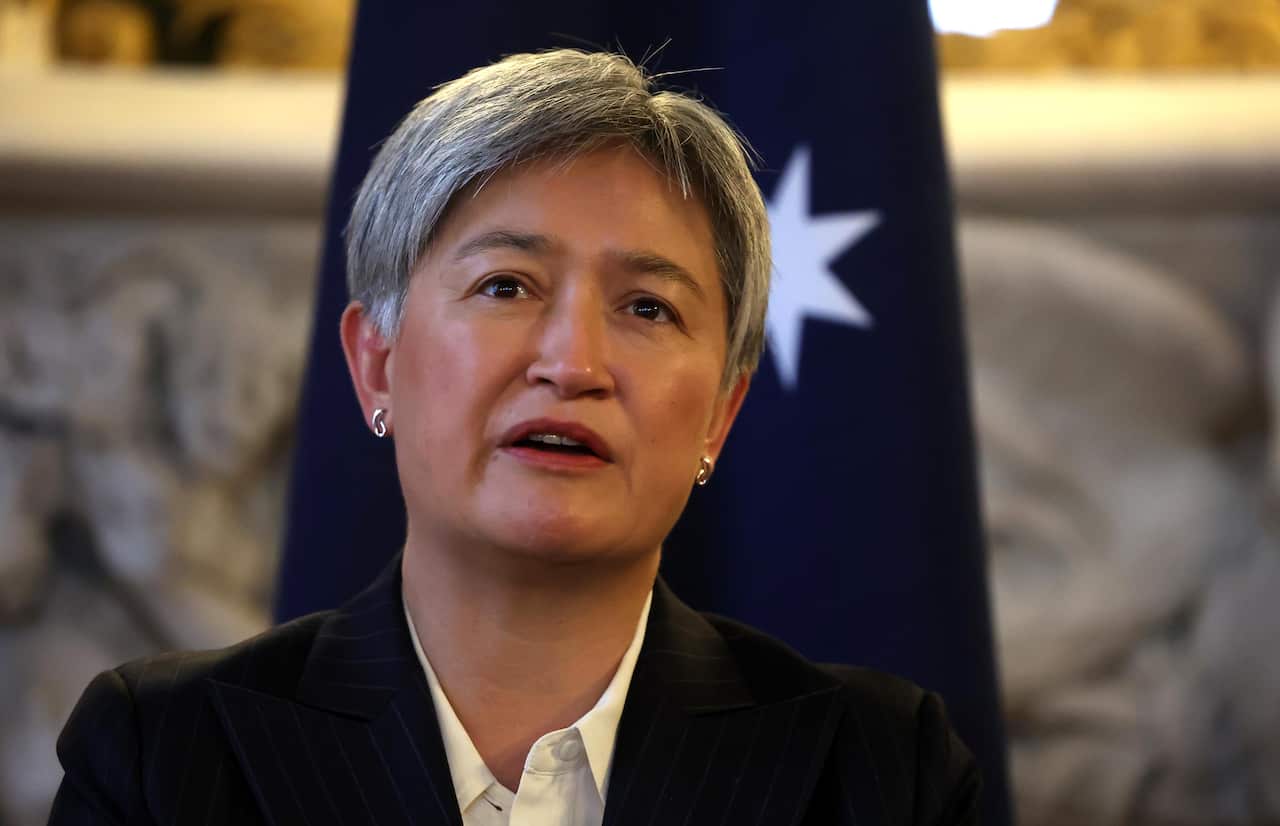 Penny Wong in a black suit and white shirt, standing in front of an Australian flag.