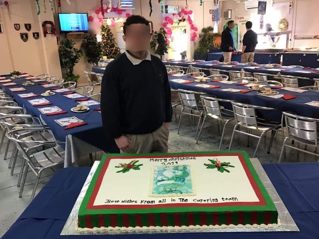 A man standing inside. A large square cake is on a table in front of him.