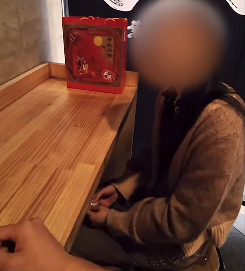 A woman with her face blurred sits at a wooden table eating from a bowl of ramen
