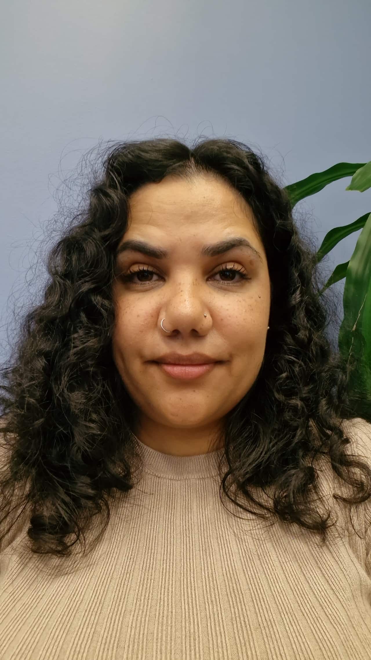 A selfie of a woman with long dark hair in a beige top. She has a slight smile on her face.