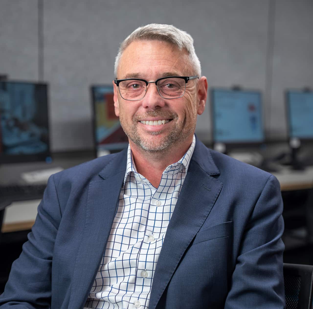 A man wearing glasses and a blue jacket sits in an office, smiling at the camera.