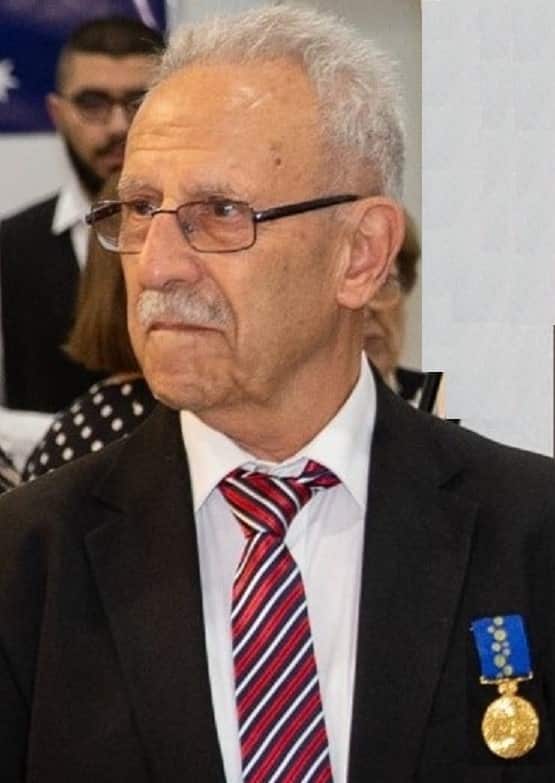 A man wearing a black jacket, white shirt and red, black and white tie, with a blue and gold medal pinned to his jacket