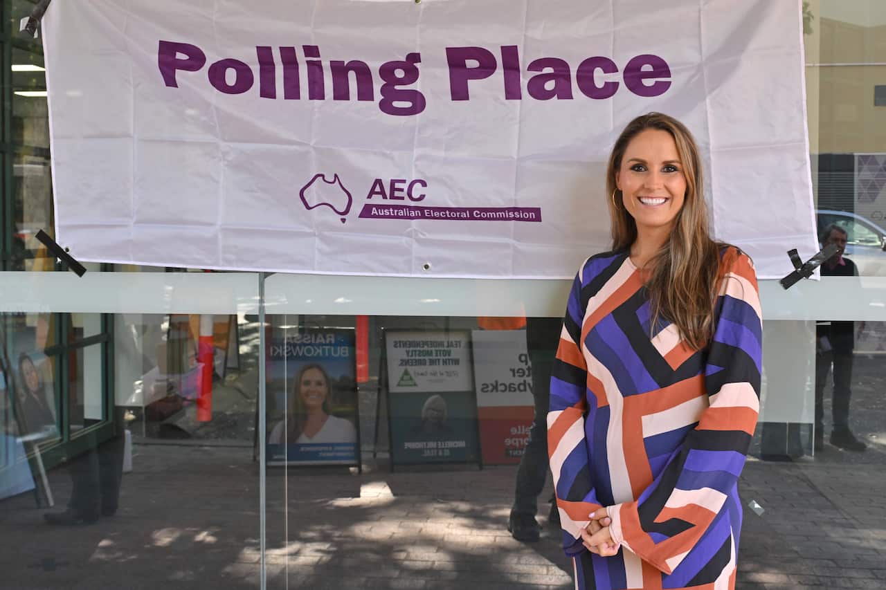 A smiling woman wearing a long-sleeved dress with a brown, blue, black and white striped pattern stands in front of an Australian Electoral Commission polling place sign