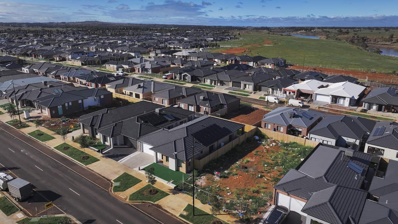 An aerial view captures a large, dense subdivision of nearly identical modern single-story houses with dark roofs. The development stretches out to the horizon, where it meets open green and brown fields under construction.