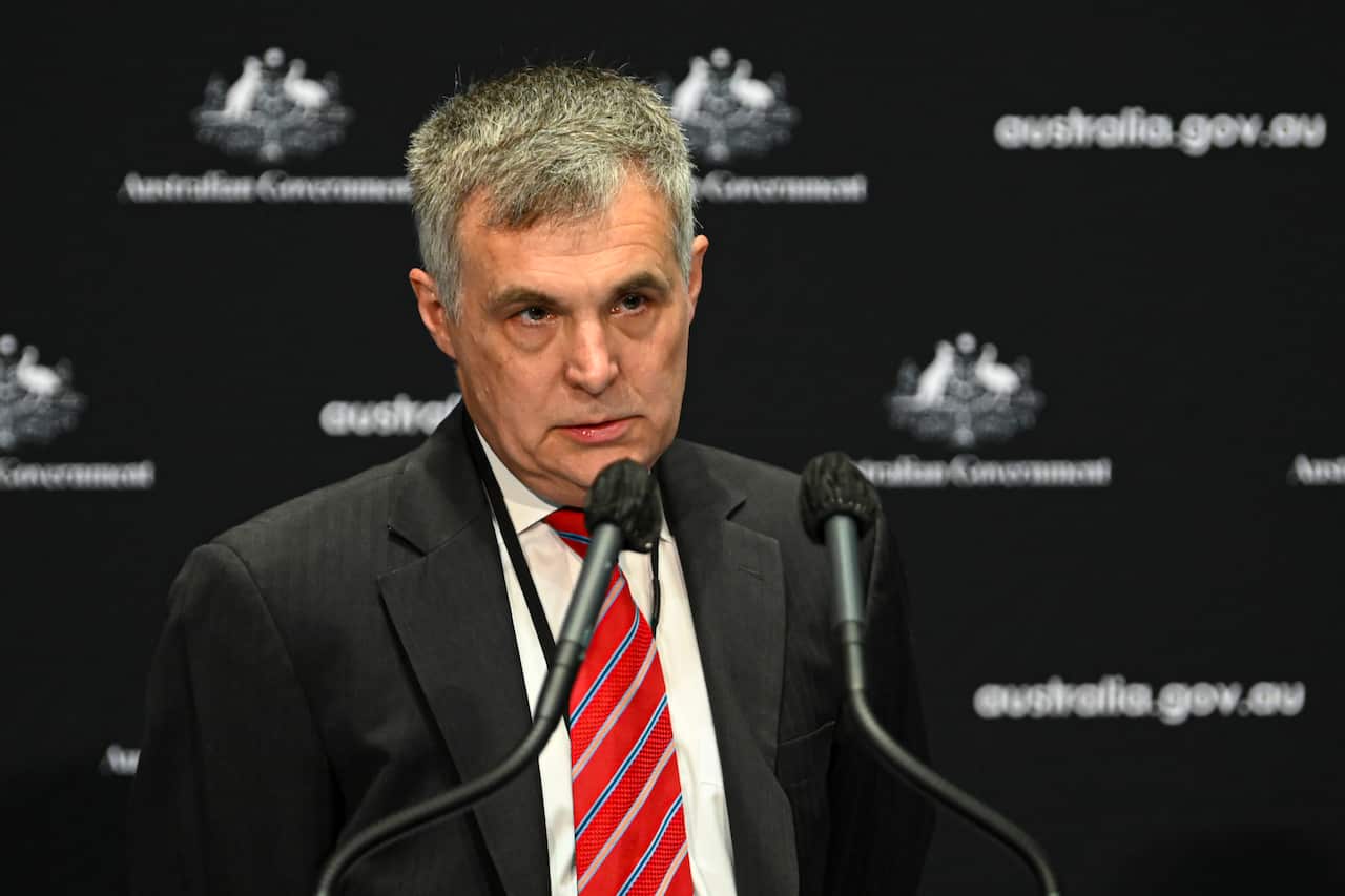 A medium close-up photograph shows a middle-aged man with grey hair wearing a dark suit, white shirt, and a red-and-white striped tie. He stands behind a microphone in front of a dark backdrop with repeating "Australian Government" and Coat of Arms logos.