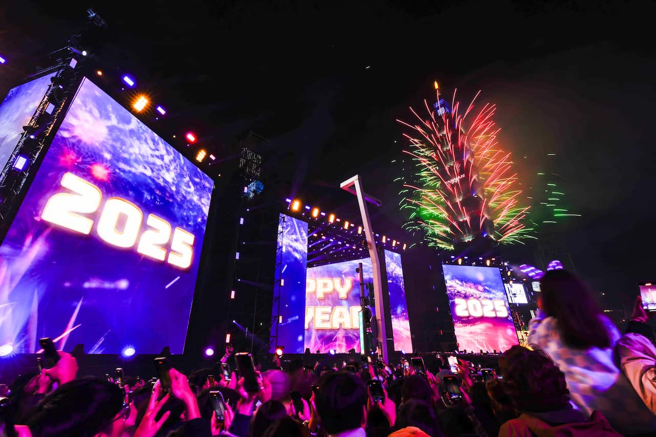 Fireworks illuminate a skyscraper as revellers watch, with other buildings featuring screens displaying fireworks and '2025'.