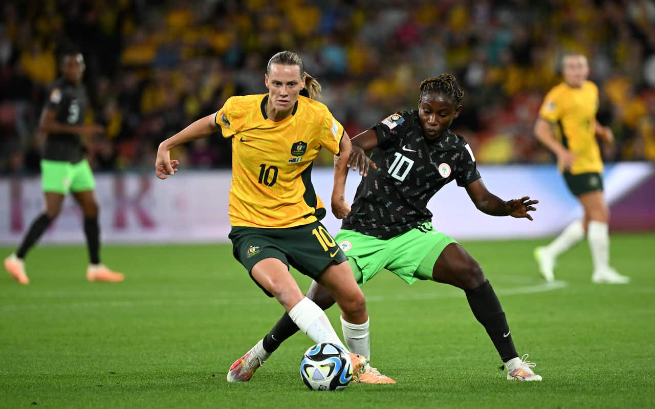 Woman in yellow and green Matildas uniform chases after soccer ball as woman in green and black uniform trails closely behind her on green pitch.