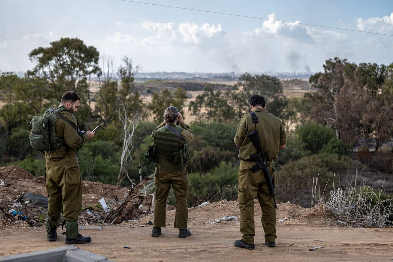 Three soldiers in military uniforms standing on a dirt road surrounded by bushland. There are smoke clouds in the distance.