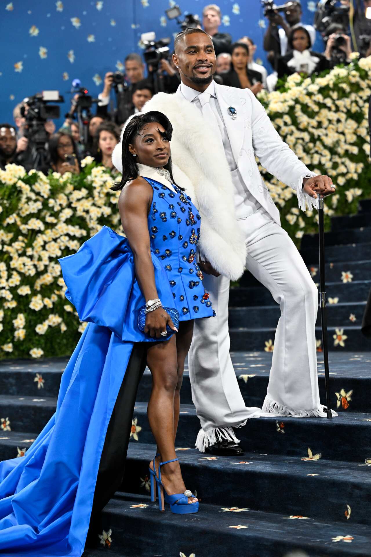 Simone Biles in a short blue dress with a long blue train stands on stairs next to a man wearing a white suit and a fur stole