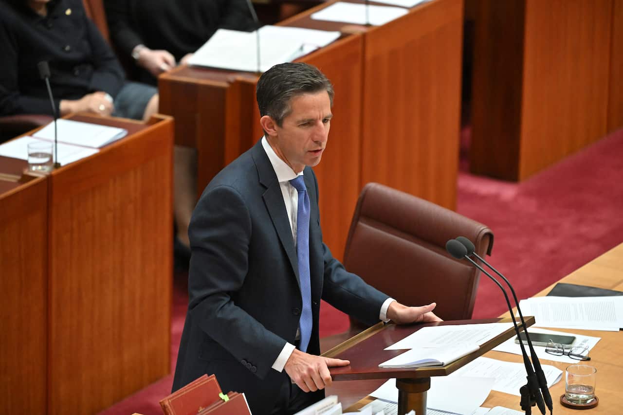 Simon Birmingham wearing a suit and tie speaking while standing in front of microphones.