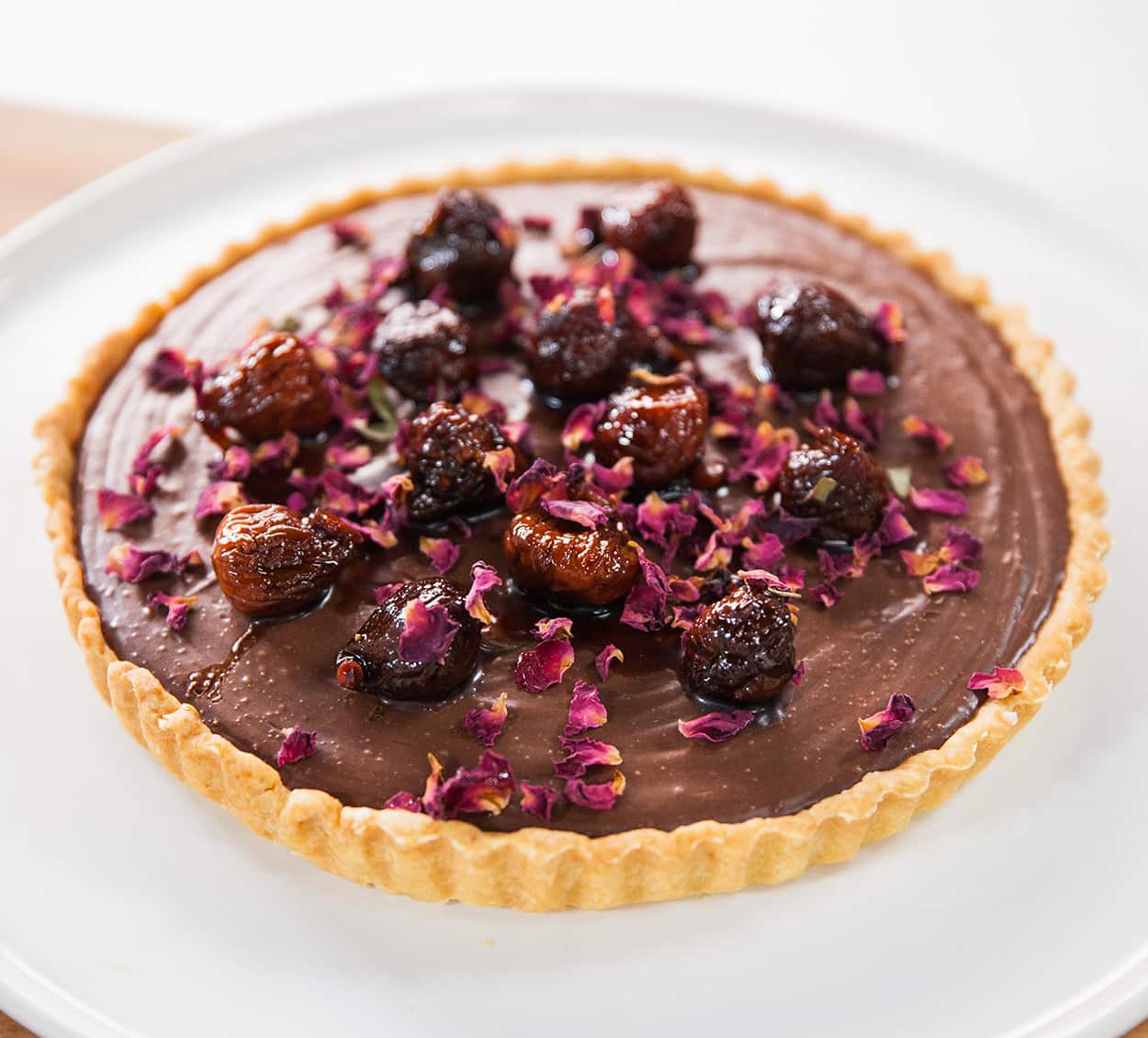 A tart with crimped pastry shell and glossy chocolate filling sites on a wide white plate. The tart is topped with cooked figs and dried rose petals. 