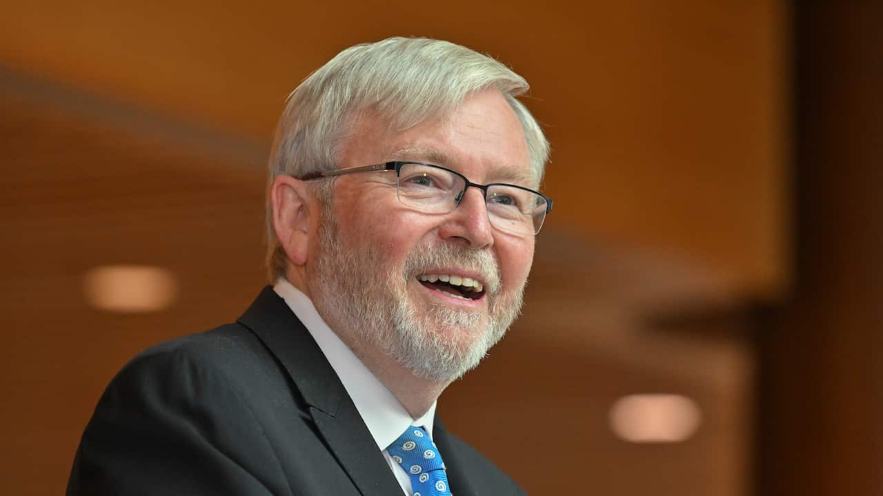 Kevin Rudd wearing a suit and smiling in front of a blurred brown wall.