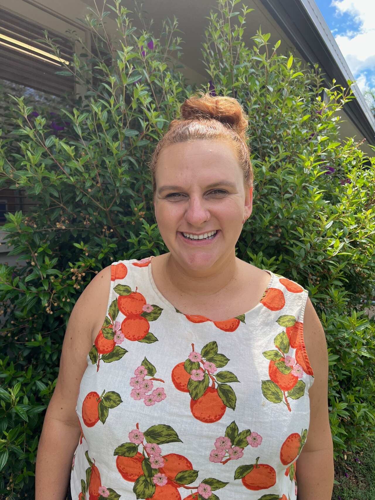 A smiling woman in a top with fruit on it, standing in front of a hedge.