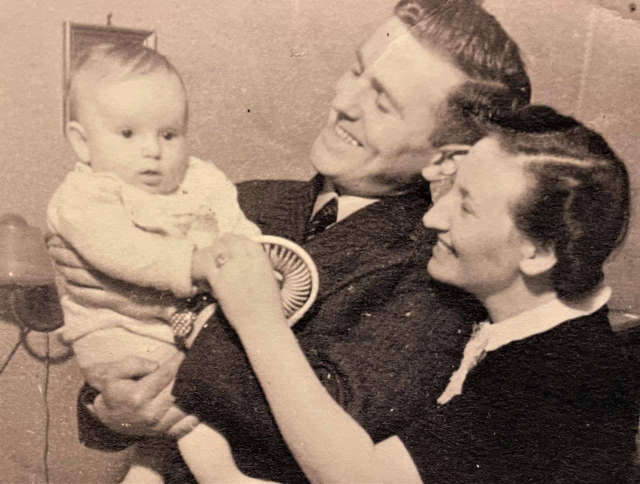 A man in a suit jacket holds a baby while a woman in a black dress holds the baby's hand.