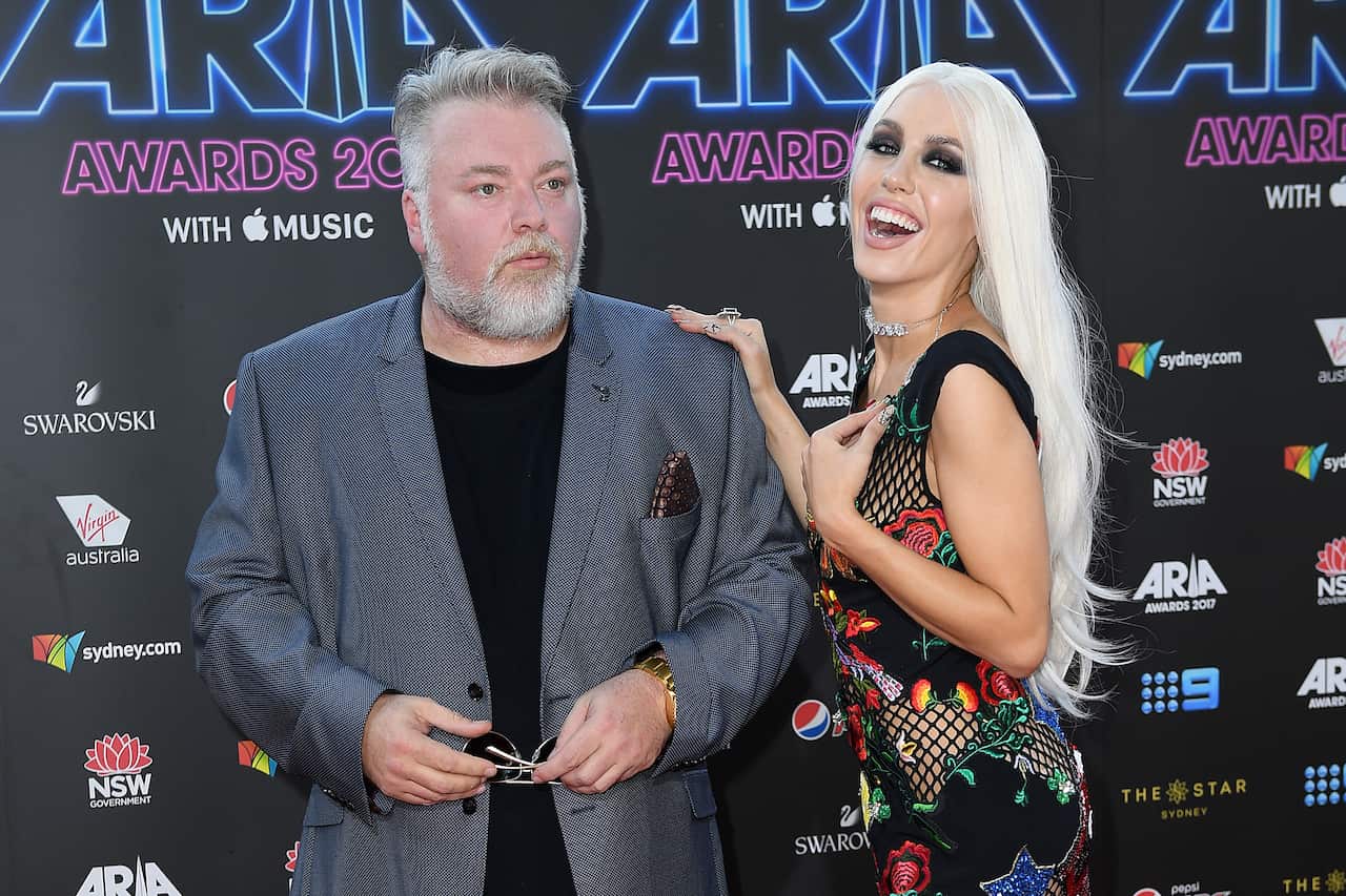 Kyle Sandilands and Imogen Anthony pose for a photo. He wears a black T-shirt and suit jacket. She is in a black dress with a colourful flower design. 