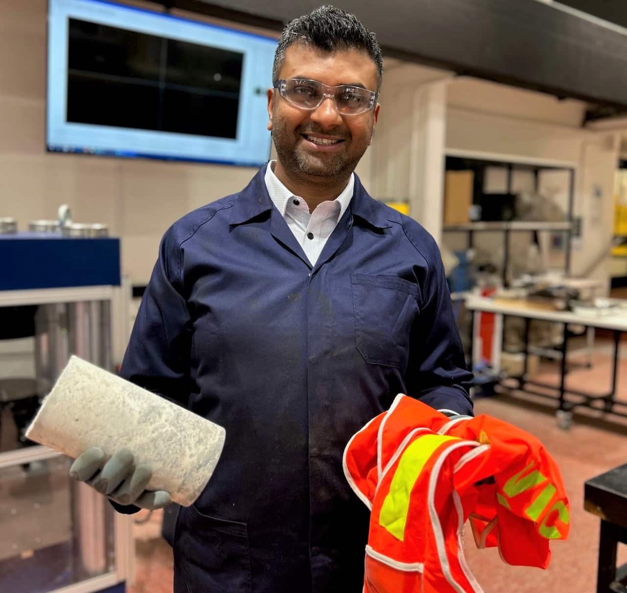 A man is smiling at the camera while holding a concrete cylinder in one hand and red hi vis vests in the other.