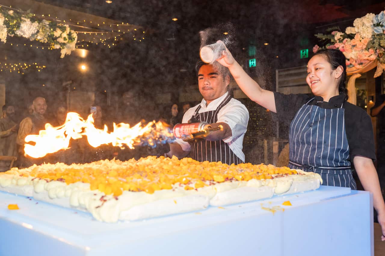 One chef uses a blowtorch to caramelise a large dessert and another chef sprinkles powder over it.