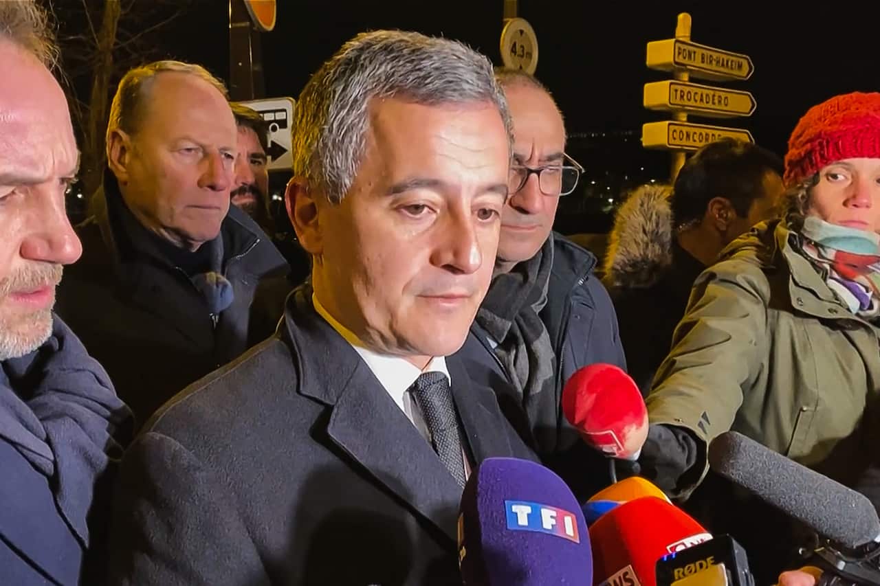 A man in a dark coat surrounded by people speaks into microphones held by reporters