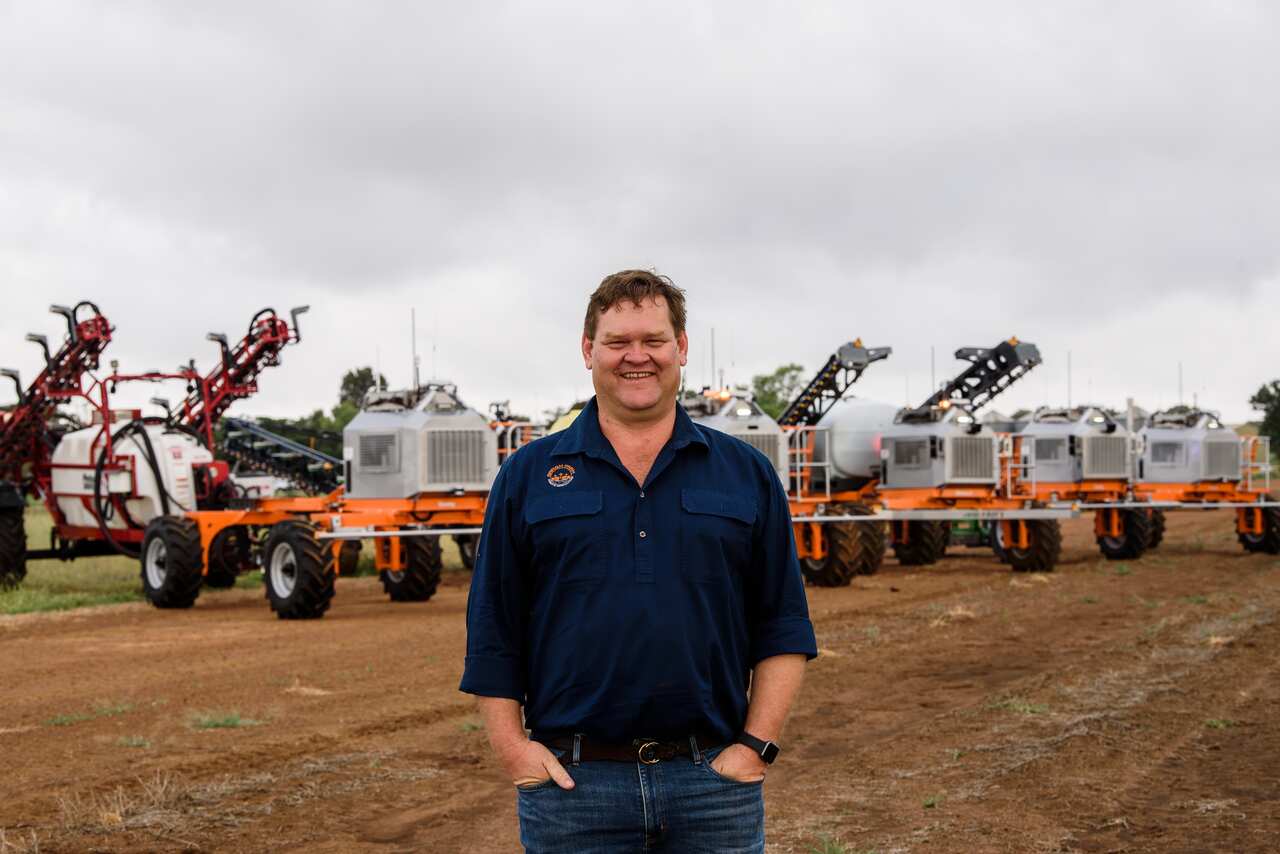 A man smiles as he stands on a field in front of a line of AI-integrated robots used for sustainable agriculture.