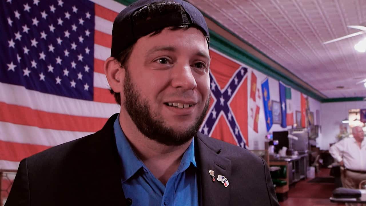 A bearded man wearing a black suit, a blue shirt, and a baseball cap backwards. In the background, there are the US flag and the Confederate battle flag.