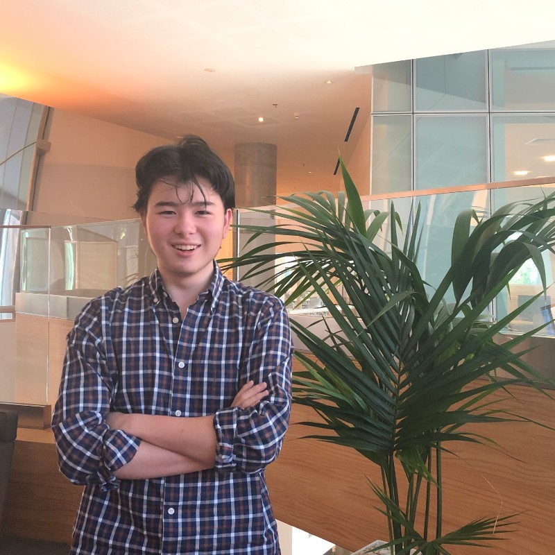 A smiling young man in a chequered shirt stands with arms folded in front of a potted plant