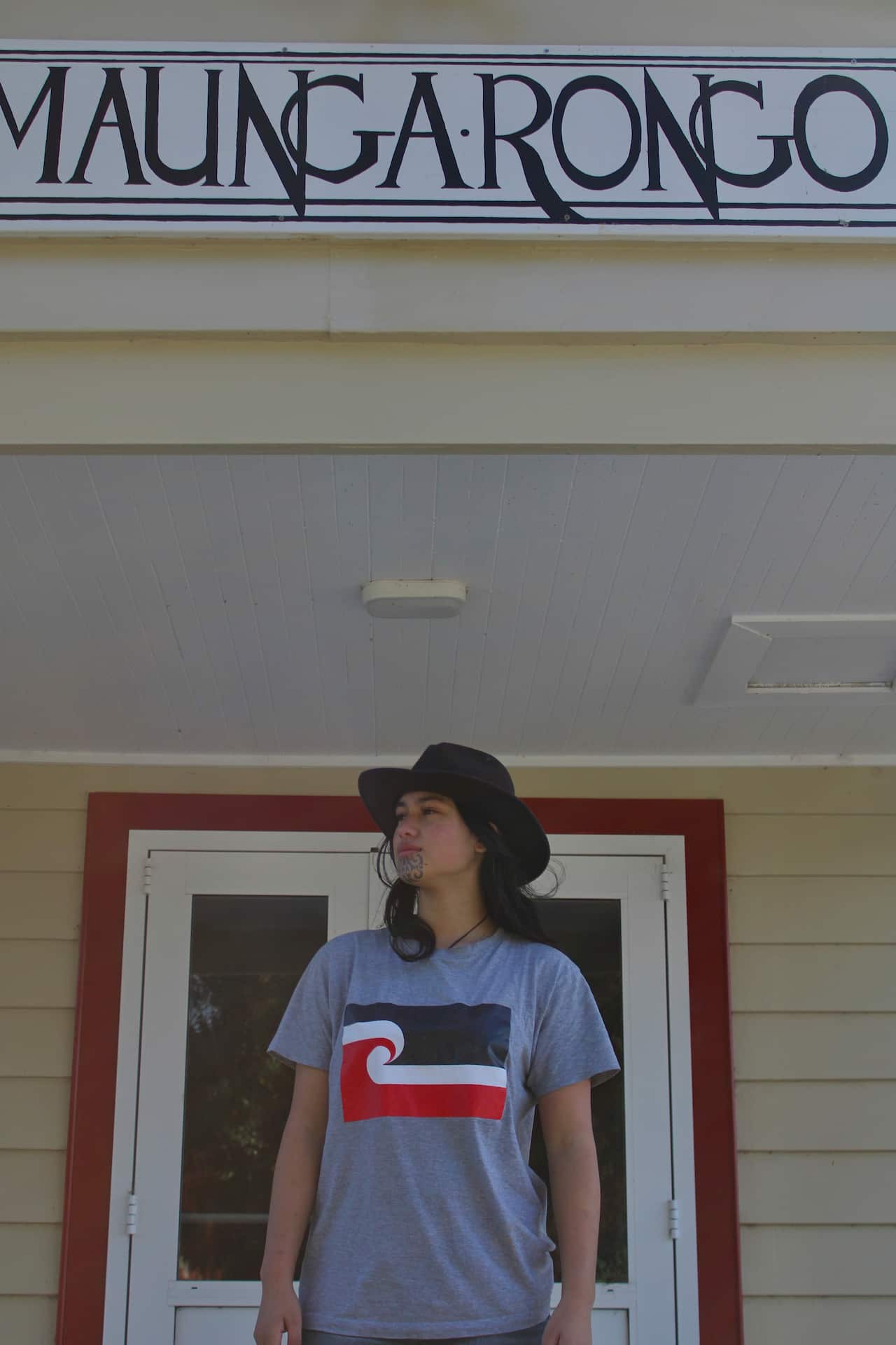 A woman wearing a grey T-shirt with the Māori flag on the front stands in front of a doorway. 