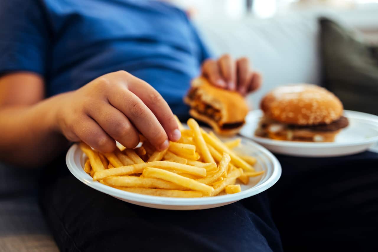 An overweight teenage boy eating a burger and chips.