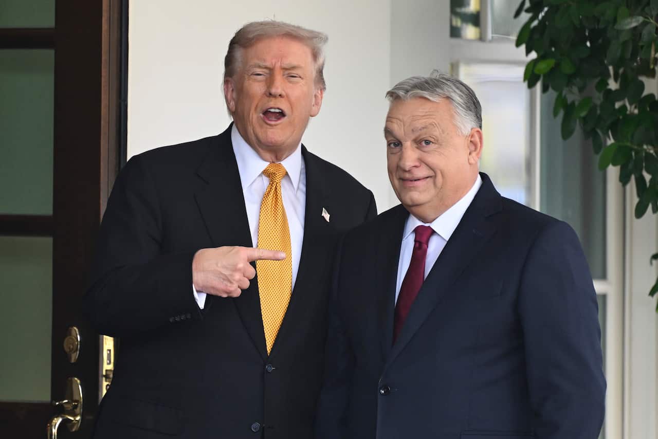 Donald Trump and Viktor Orban standing next to each other outside a white building. Trump is pointing at Orban. Both men are wearing dark suits