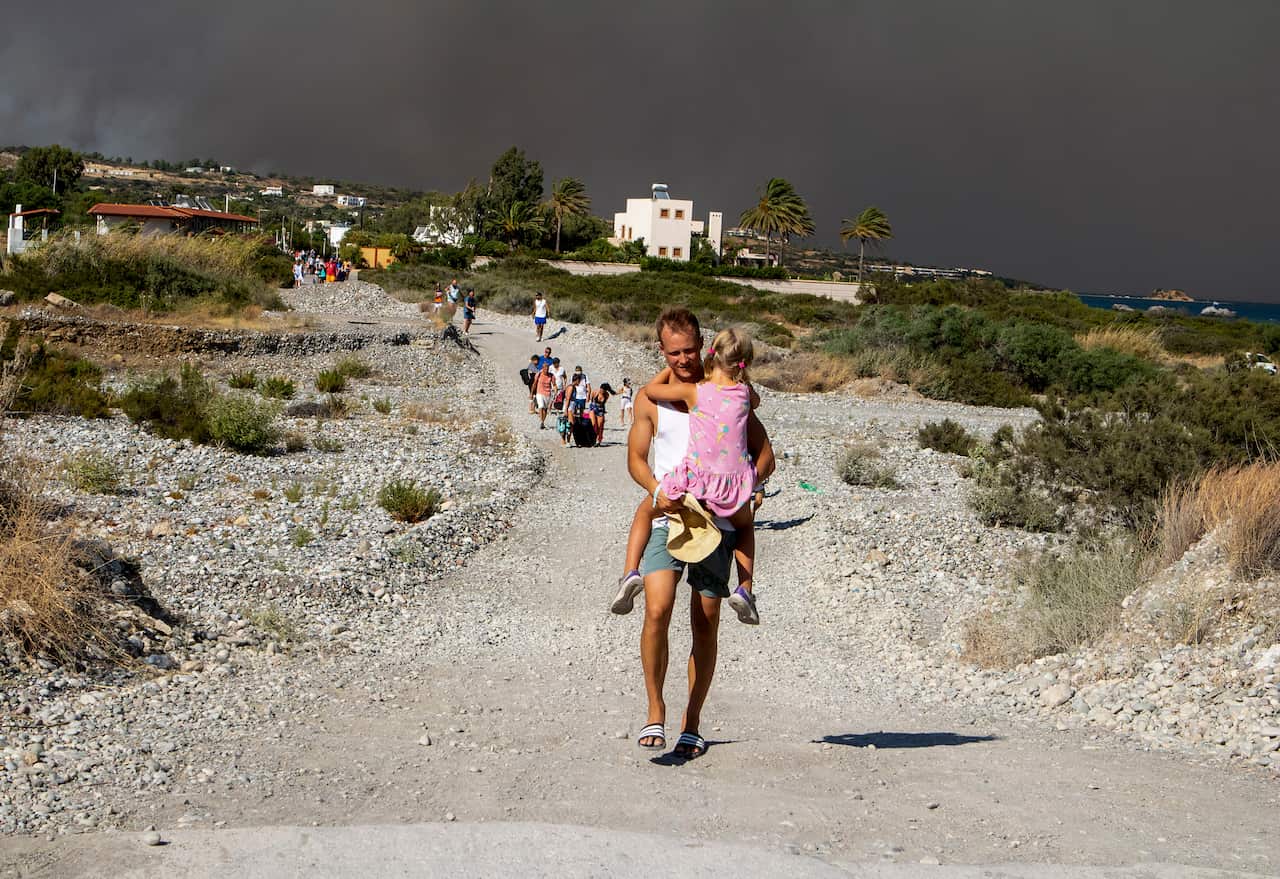 A man in shorts carries a young girl while he walks down a hill, with a smoky sky in the background. 