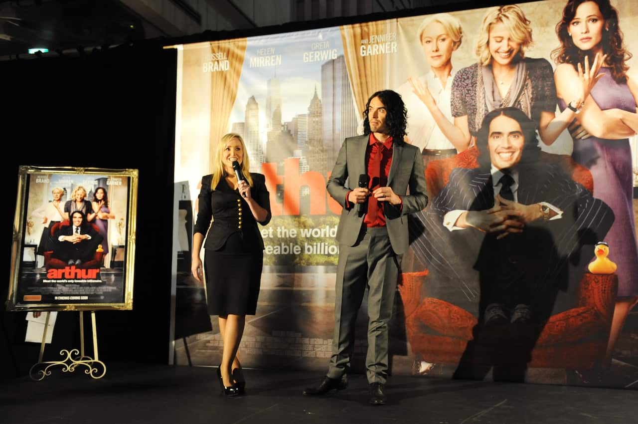 Russell Brand and Fifi Box at the premiere of his film Arthur. Fifi Box is wearing a black shirt and skirt, and Russell Brand is wearing a grey suit and red shirt. They are both holding microphones. 