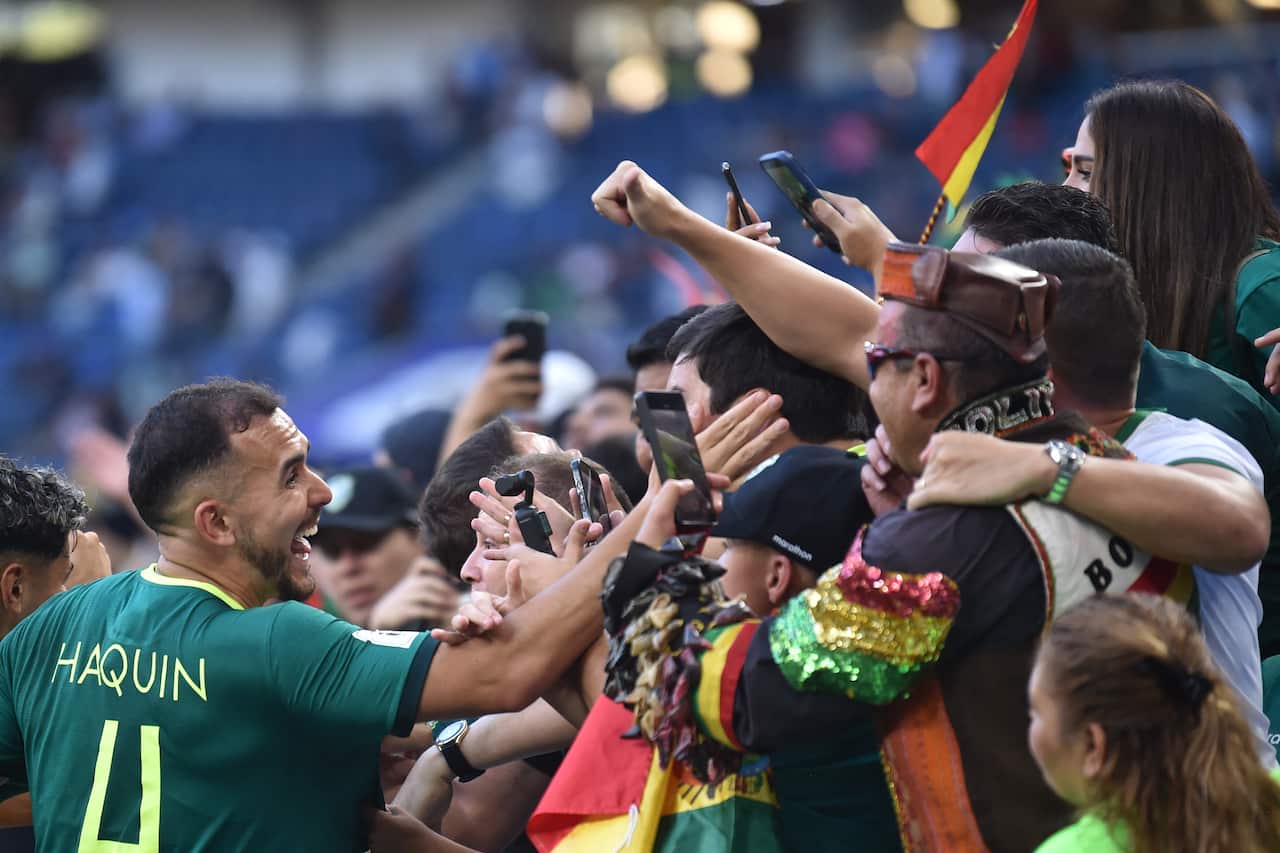 Bilivian football player in green kit reaches out over a barrier to celebrate with happy fans waving phones and flags