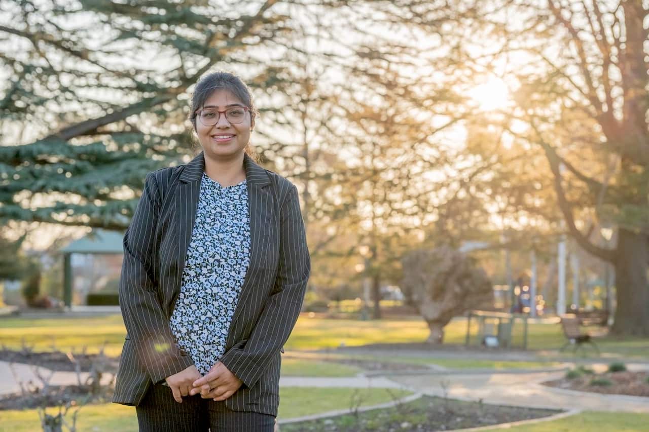 A woman in a floral shirt and pinstripe jacket stands in a park.