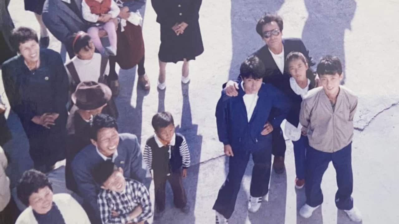 A man wearing sunglasses is looking up at the camera with his hands on the shoulders of his three teenage children. They are standing in a crowd of other people