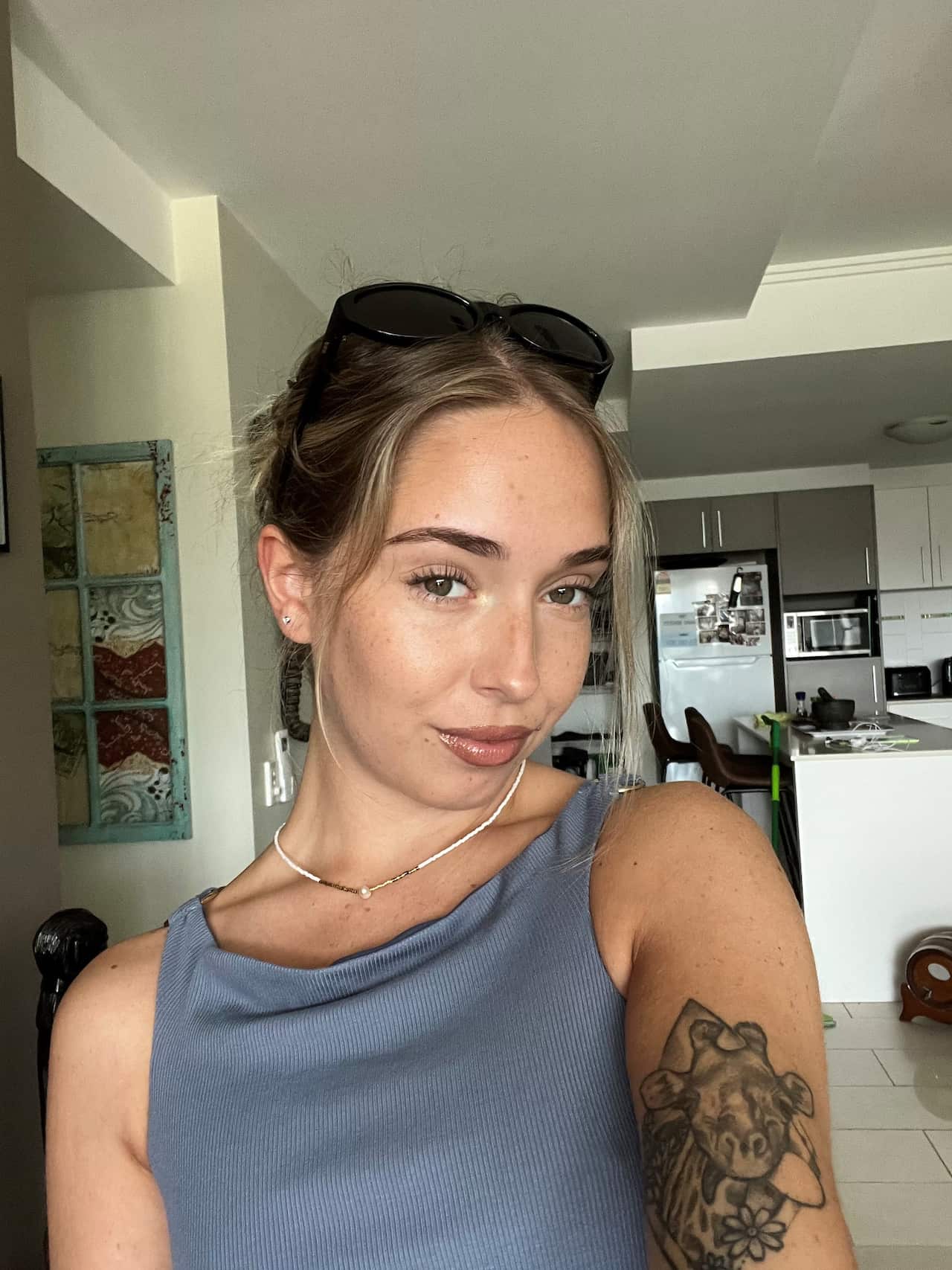 A woman with short hair, a blue top and a tattoo on her arm is taking a selfie with her kitchen in the background