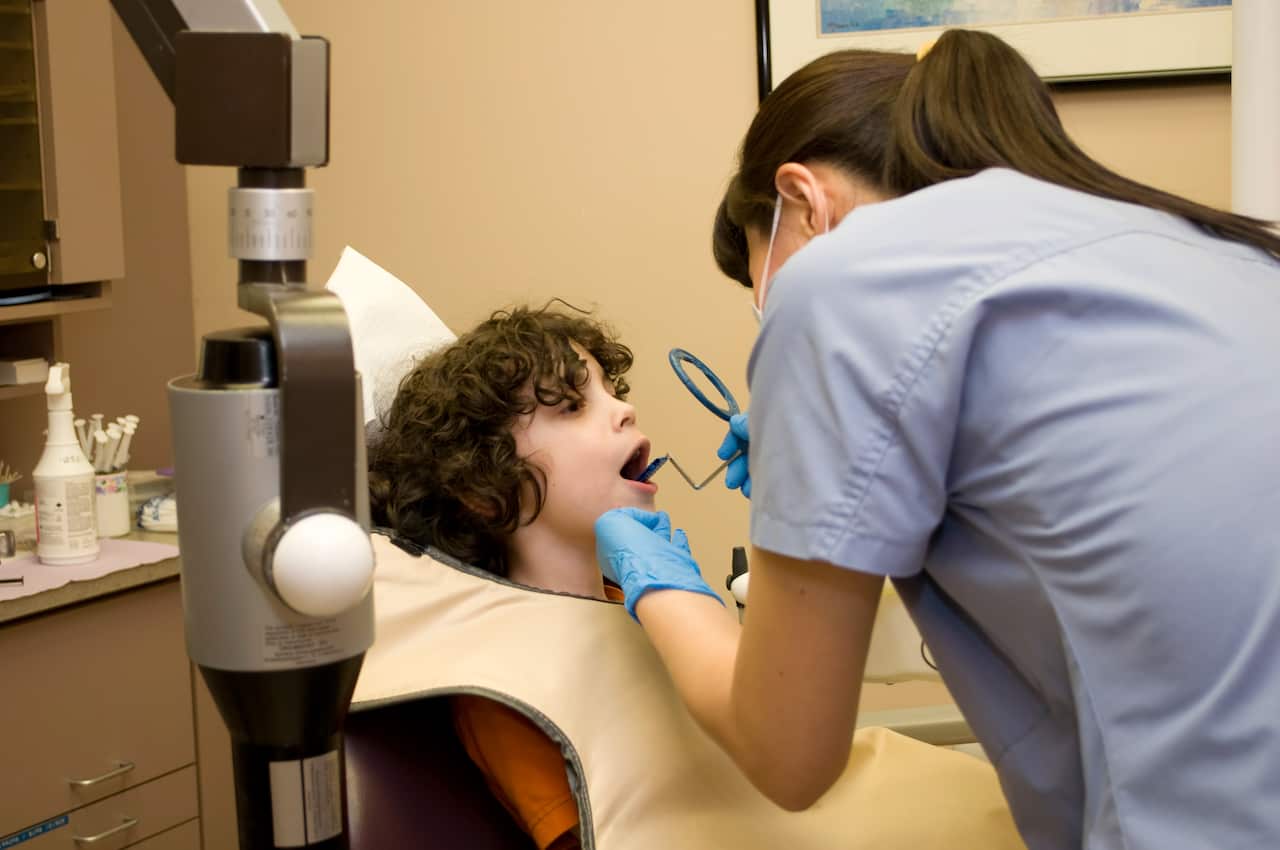 A young boy in a dentist chair while a dentist or dental technician looks into his mouth with an implement. 