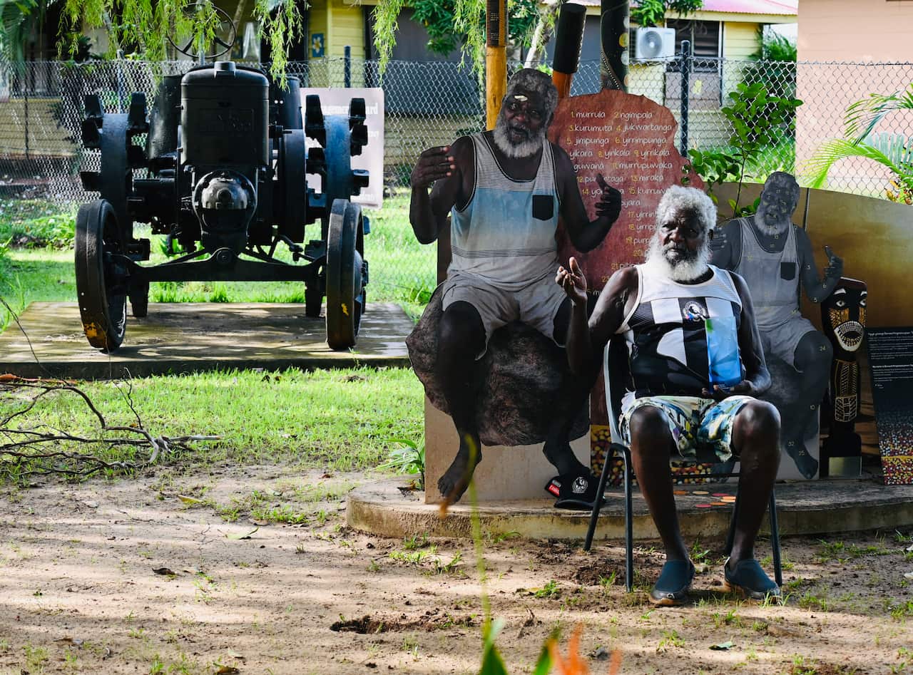 A man wearing a Collingwood Aussie Rules jumper sitting in a chair outside in front of a life-size cut-out of himself.
