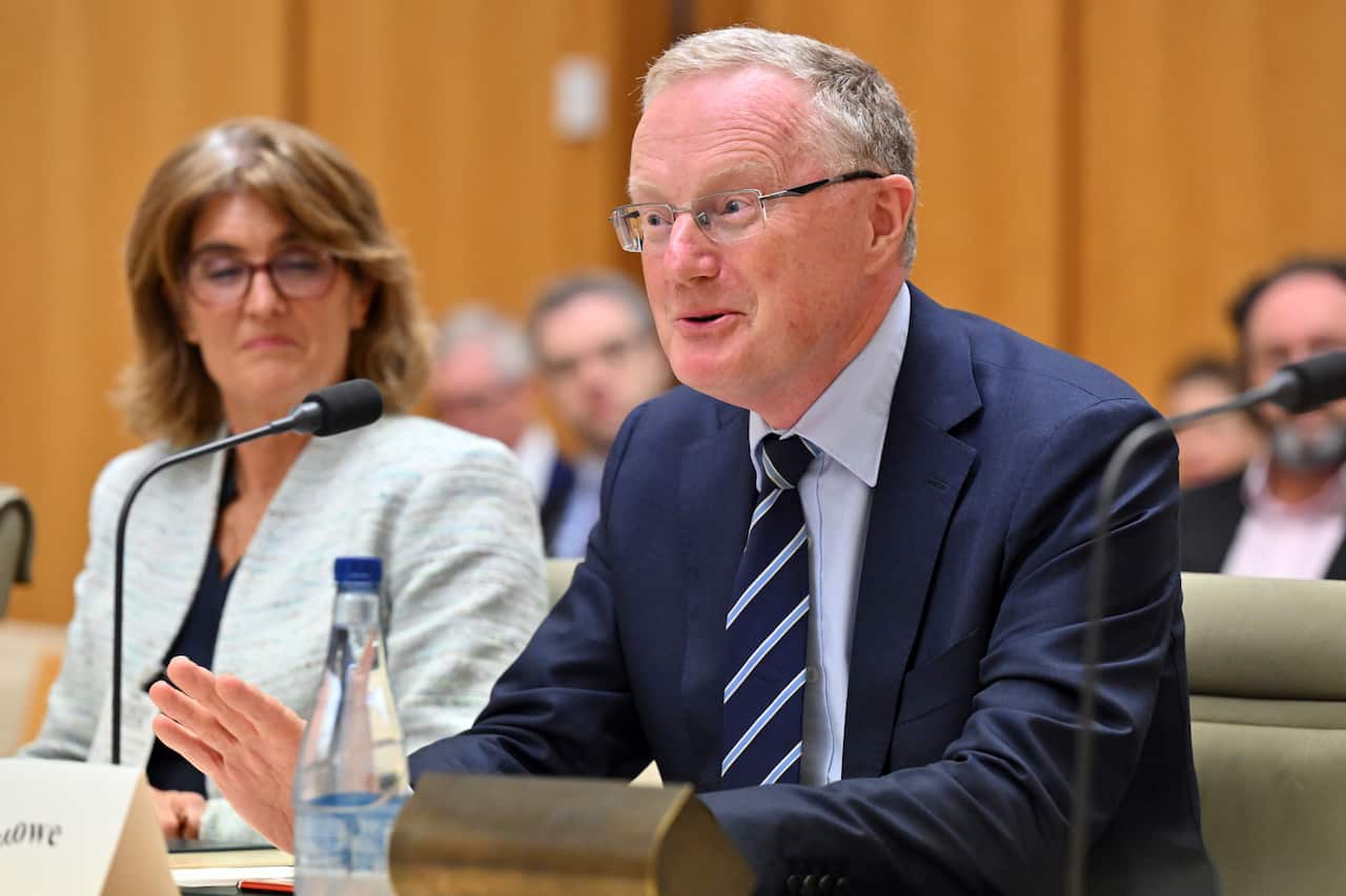A man wearing a suit, tie and glasses seated at a table and speaking in front of a microphone.