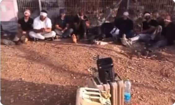 A group of men blindfolded and sat on the ground with a speaker in front of them