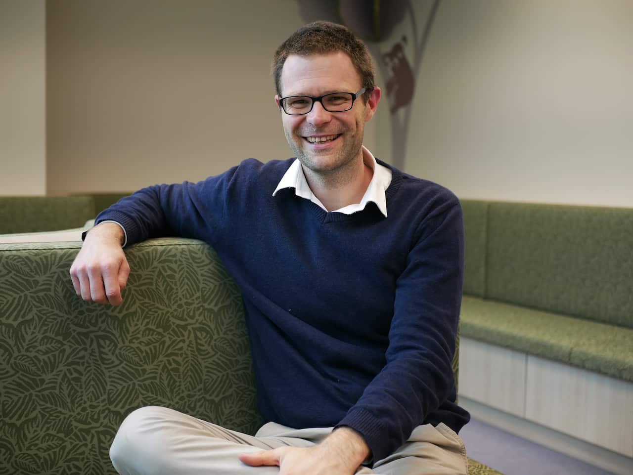 A man wearing light-coloured trousers and a blue jumper sitting down facing the camera