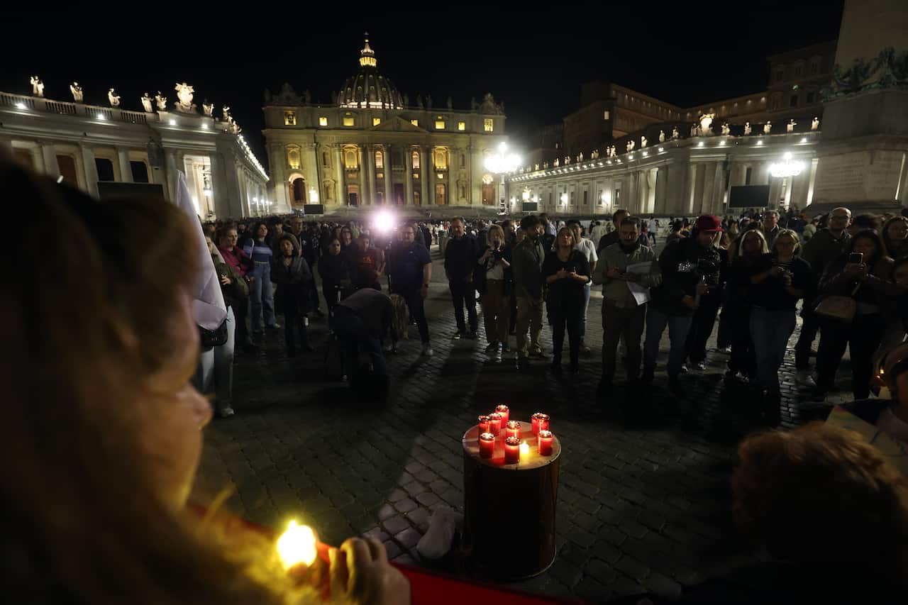 People light candles at a square outside a well-lit white building at night.