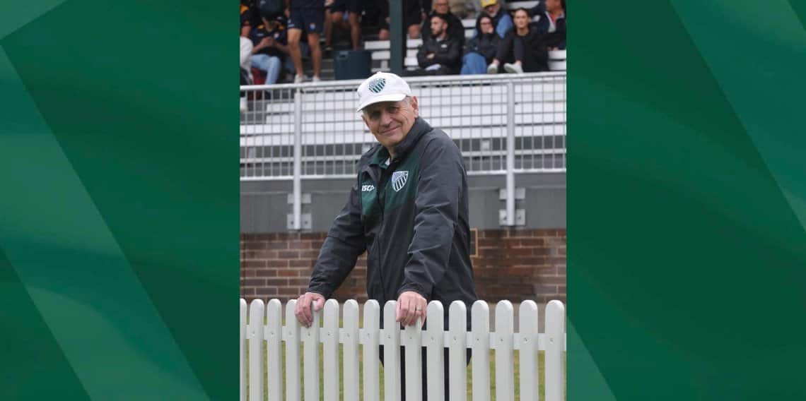 A man wearing a sports cap and jacket smiles while leaning on a short white fence. 
