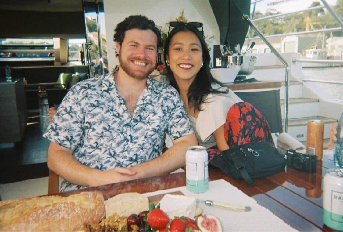 A man in a blue and white short-sleeved shirt sits close to a woman at a table. There is a cheese and fruit platter on the table in front of them