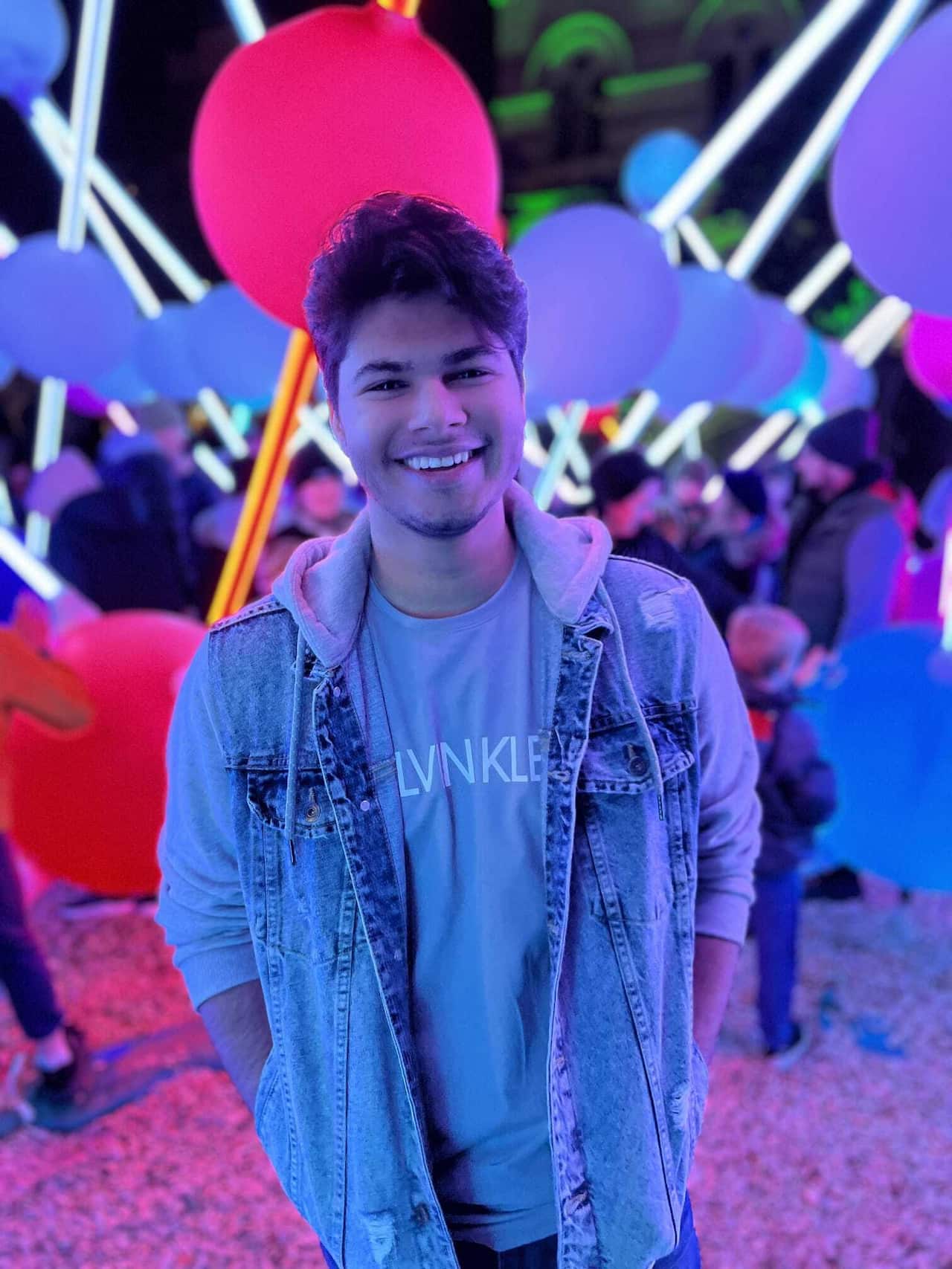 Tejas Gandhi smiles while looking directly at the camera. He is wearing a long sleeve shirt and denim vest, and is lit up by bright multicoloured lights behind him. 