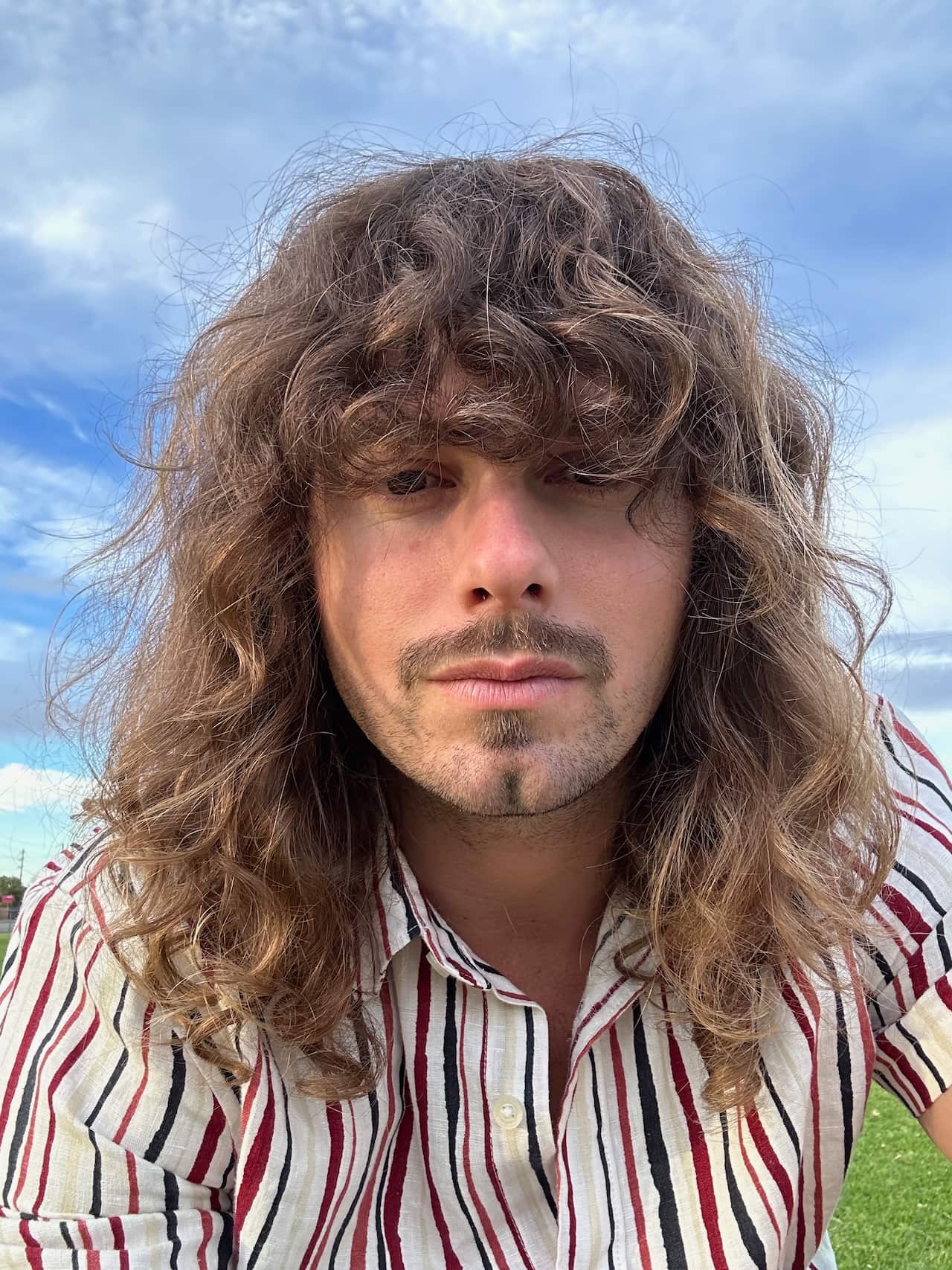 A close-up photo of a man with long, curly hair wearing a striped collared shirt